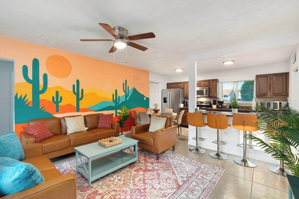 Bright living room with a colorful desert mural, cozy sofa seating, patterned rug, and ceiling fan flowing into the kitchen with bar stools—an inviting space to gather, chat, and relax together indoors.
