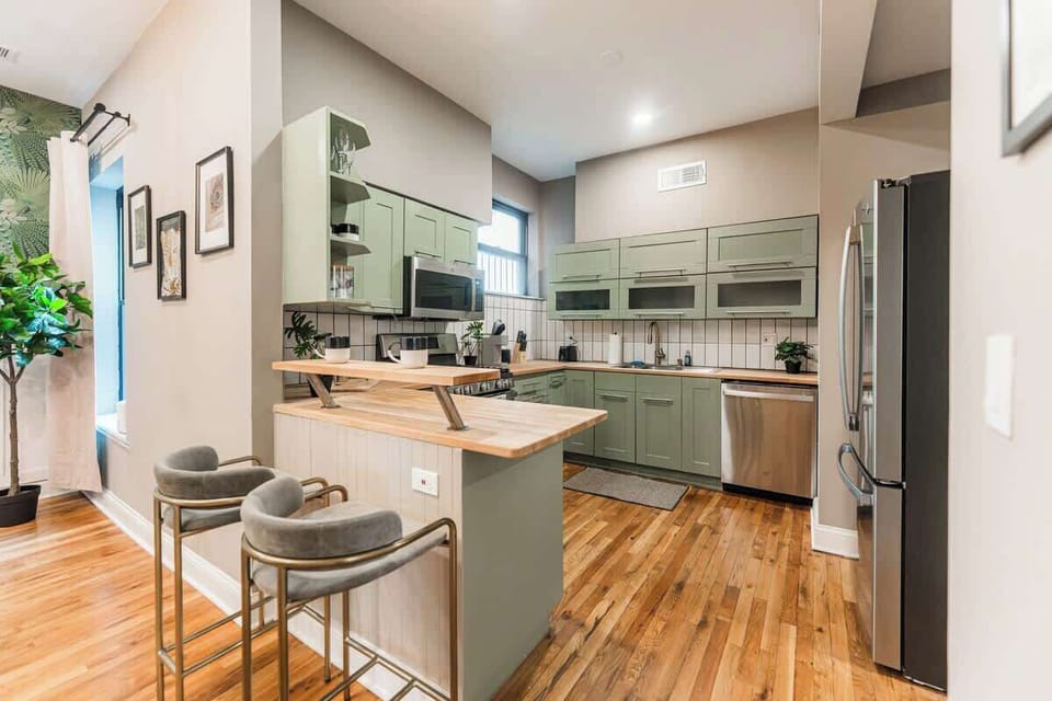 Spacious kitchen with sage green cabinets, breakfast bar seating, and stainless steel appliances. Open layout with warm wood floors—perfect for cooking, dining, and socializing.
