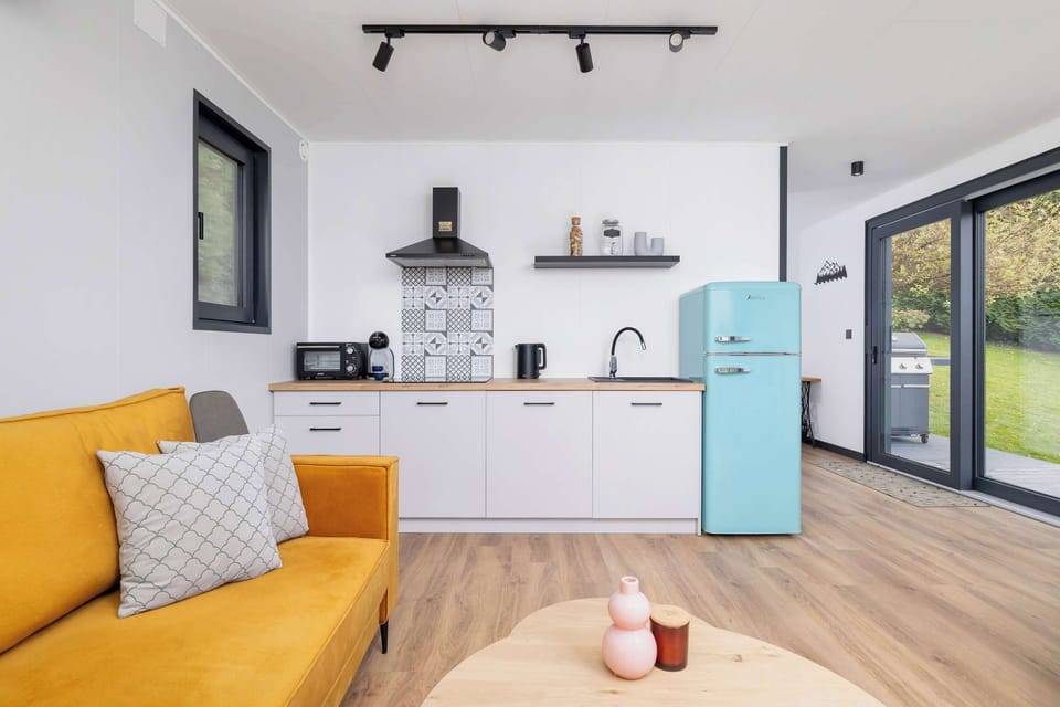 A bright open-plan living room with a white kitchen, yellow sofa, and pastel-blue refrigerator. The sliding doors open to a terrace with a garden view.