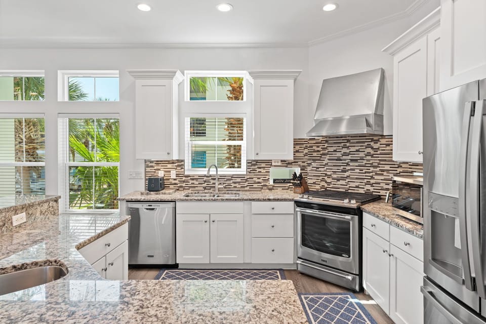 Bright coastal kitchen featuring granite countertops, stainless steel appliances, and a mosaic backsplash. Sunlit windows and crisp white cabinetry create an inviting seaside atmosphere.