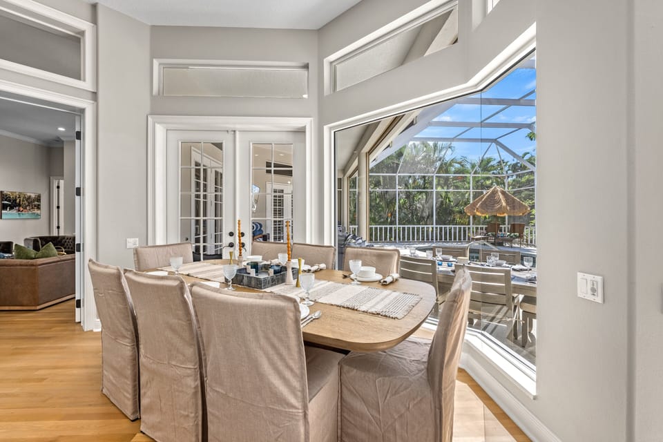 The dining space is bathed in natural light, with full glass doors framing the sparkling pool outside.
