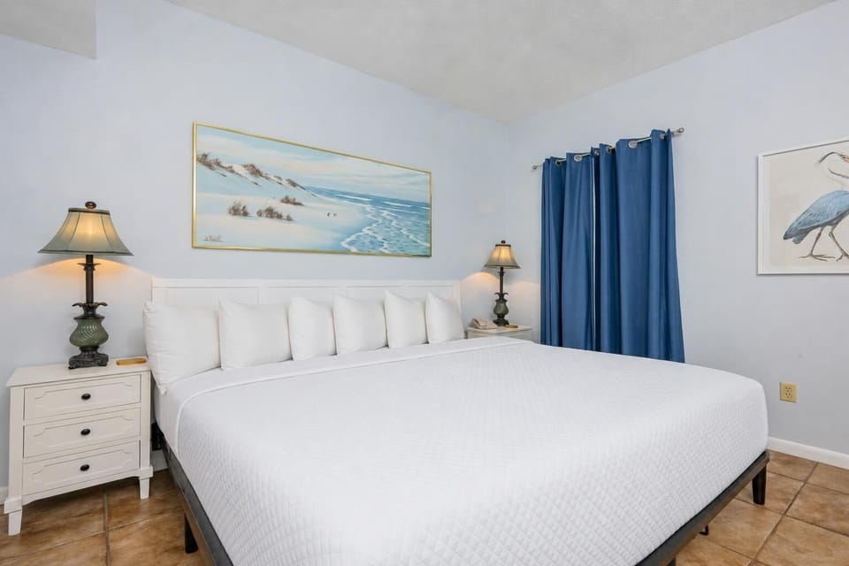 This cozy bedroom features a large king bed with white linens, charming lamps, and vibrant blue curtains