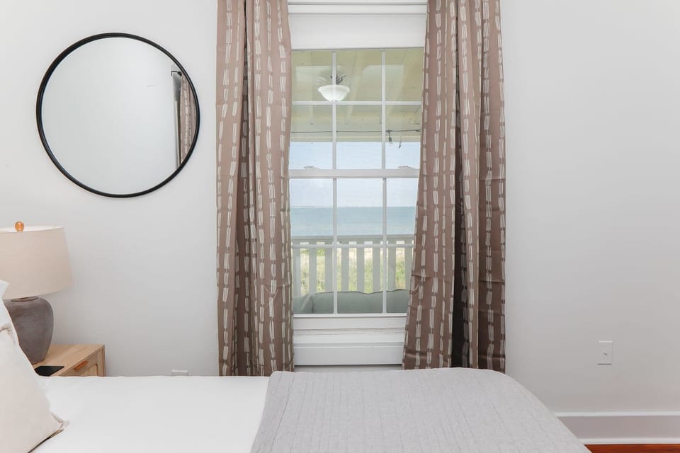 Natural light pours in through the window, highlighting the calming neutral tones and elegant decor. A bedside lamp and blackout curtains ensure a perfect night's rest.