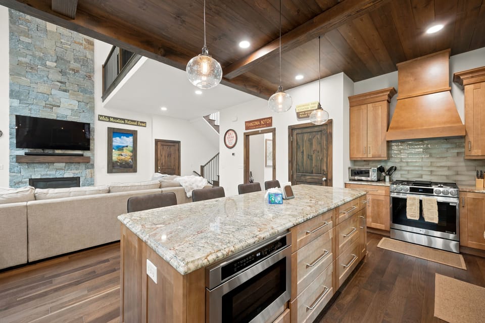 Open-concept kitchen and living area with a large granite island and stone fireplace. Perfect for entertaining, cooking, and relaxing together in a warm, inviting space.