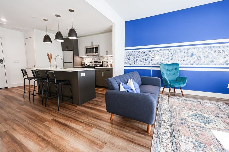 A contemporary inviting studio, you'll find a well-appointed kitchen with bar seating, a cozy living area adorned with chic furniture, highlighted by a striking blue wall and ample natural light