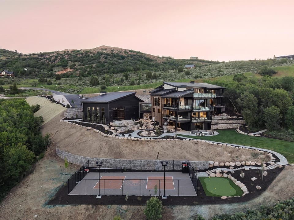 An expansive estate nestled in rolling hills, featuring modern architecture, landscaped grounds, a tennis court, and mini golf, all set against a dramatic natural backdrop.