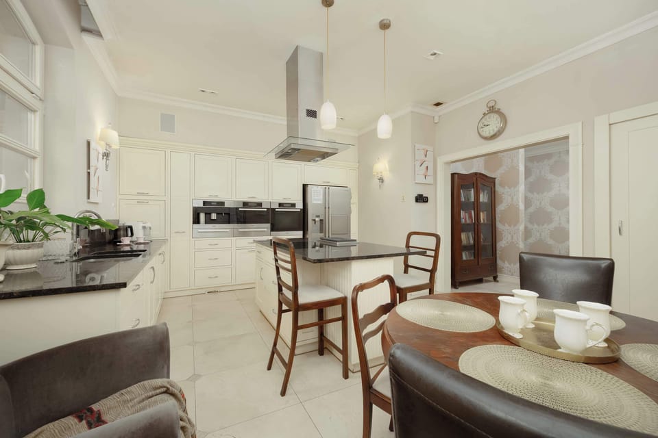 A bright kitchen with elegant white cabinetry, modern appliances, and a breakfast area. The room is filled with natural light from large windows.
