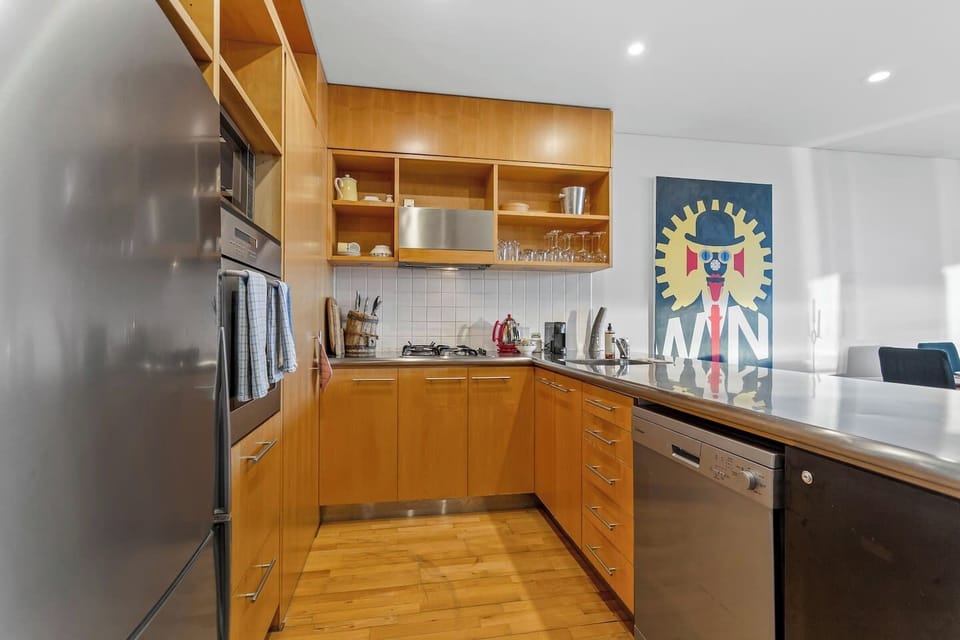 The kitchen from a different angle — U-shaped bench run, dishwasher, oven, gas hob, and open shelving stocked with everything you could need. Designed for people who actually cook