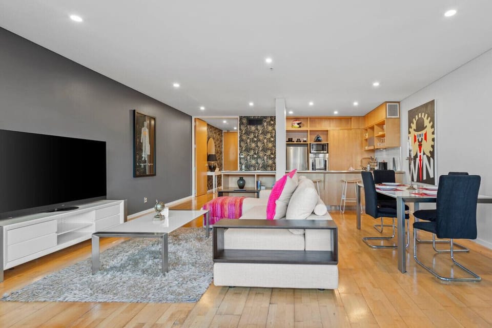 A living space that feels alive — bold artwork, warm timber floors, a generous sofa dressed with pink and magenta cushions, and a kitchen that says someone actually cooks here