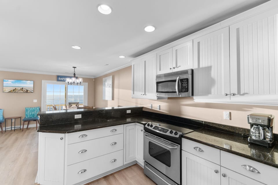 Step into your fully-equipped kitchen featuring granite countertops, stainless steel appliances, and bright coastal views through the window.