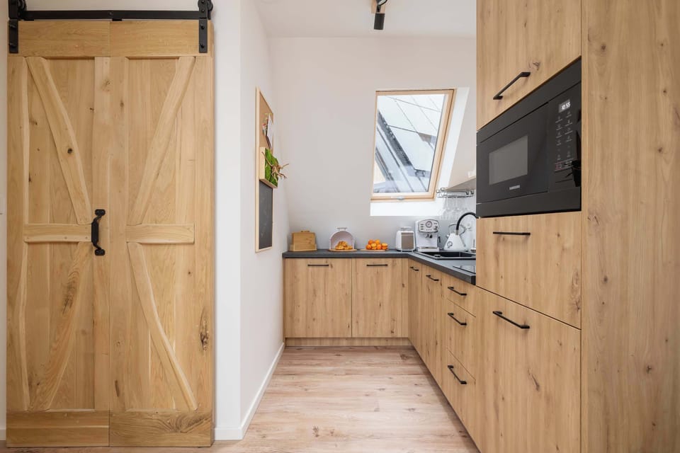 A small modern kitchen under slanted ceilings with a window bringing in daylight. The combination of wood and black fixtures creates contrast.