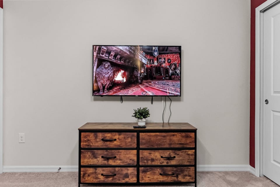 Enjoy a cozy atmosphere with this warm-toned wooden dresser and sleek flat-screen TV above. The soft wall color provides a serene backdrop, while the simple greenery adds a lively touch to your space.