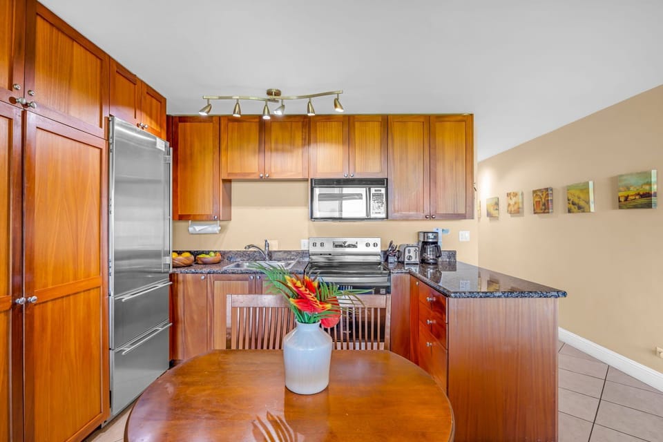 Enjoy a well-appointed kitchen with rich wood cabinetry, sleek granite countertops, and modern stainless-steel appliances, complemented by a cozy dining table with a fresh floral accent.