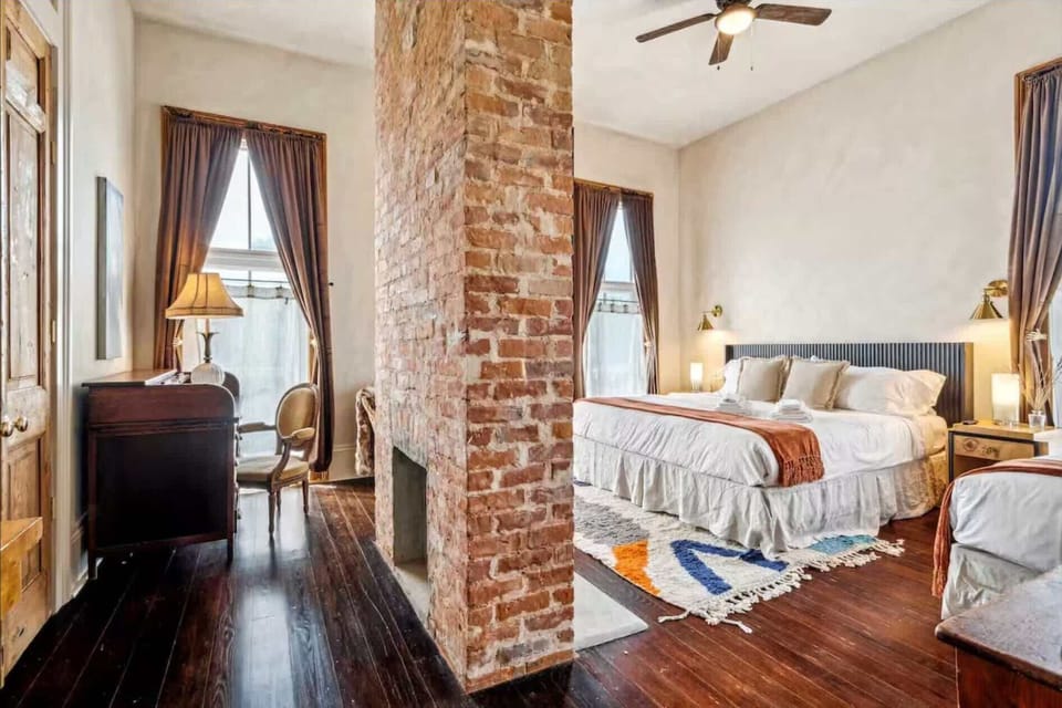 Spacious king suite with exposed brick, vintage desk nook, and warm natural light throughout.
The large front windows highlight the cozy seating area and restored historic fireplace.