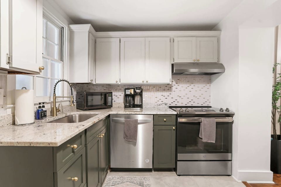 The fully equipped kitchen features sage green cabinetry with brass hardware, granite countertops, a patterned tile backsplash, stainless steel appliances, and plenty of prep space for cooking in.