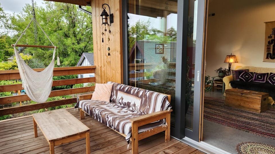 Covered balcony with wood plank flooring, hanging macrame chair, patterned throw on wooden loveseat, and sliding glass door.