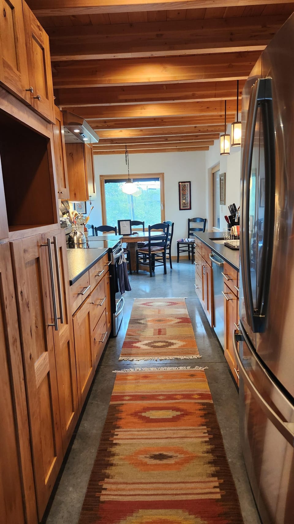 Narrow kitchen with exposed wood beams, stainless steel appliances, black countertops, and patterned runners on concrete floor.