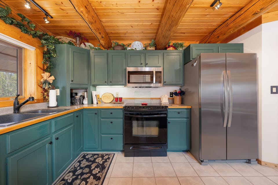 Your rustic kitchen blends cozy cabin charm with modern convenience, featuring sage-green cabinets and warm wood beams overhead.