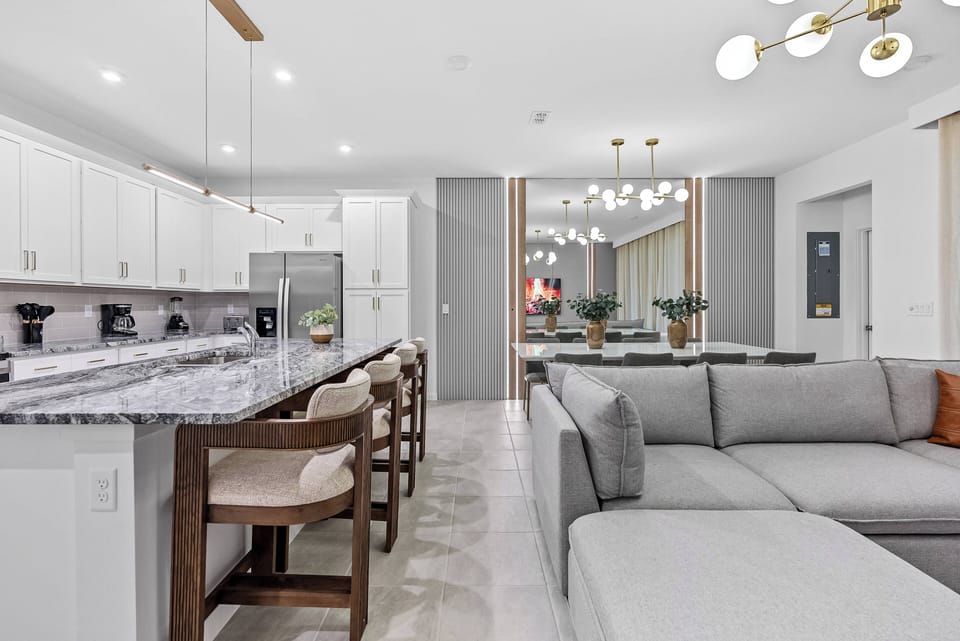 Relax in the spacious living area with a sleek gray sectional and light wood bar stools at the kitchen island. Enjoy the natural light illuminating the elegant granite countertops and modern lighting for a chic, cozy atmosphere.