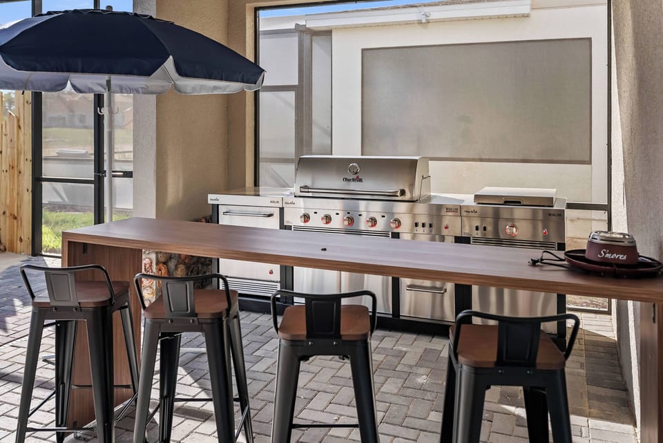 Experience a chic outdoor kitchen with modern stainless steel appliances and a bar-height table, perfect for casual dining. Enjoy the natural light and shaded comfort of an umbrella, ideal for gatherings and delightful meals.