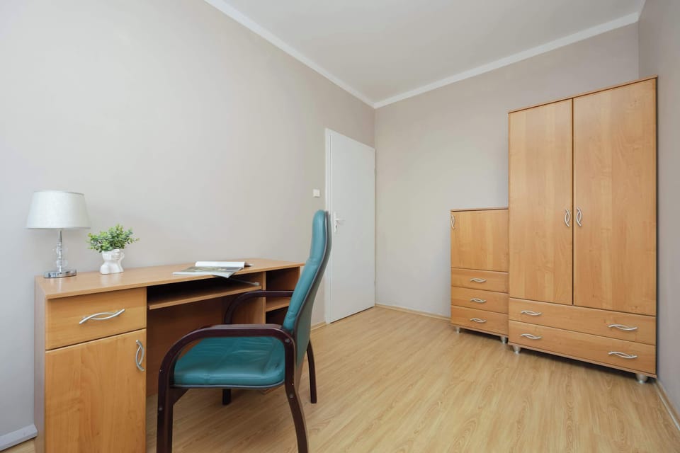 A bright room with a double bed and wooden wardrobe. A green chair sits beside a writing desk by the window.