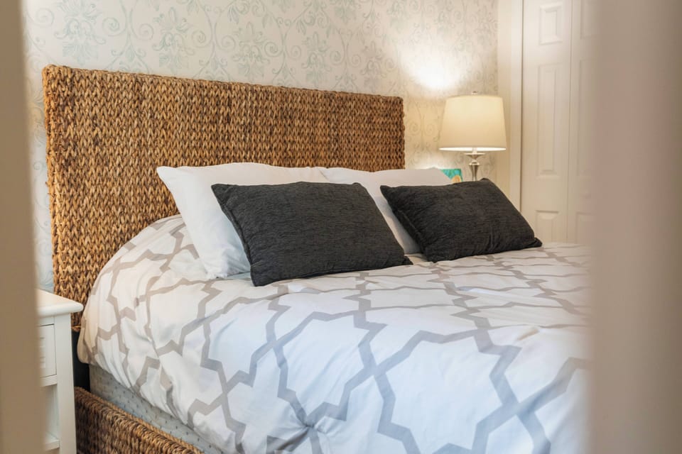 Experience restful nights in this cozy bedroom featuring a warm woven headboard. The soft patterned bedding and serene colors create a tranquil atmosphere, while the bedside lamp offers a gentle glow for your relaxing evenings.