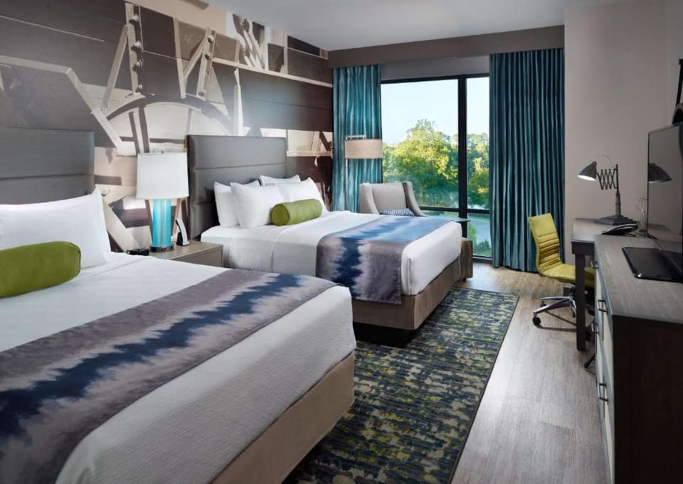 Two beds with bright decor, large window, and work desk by the view