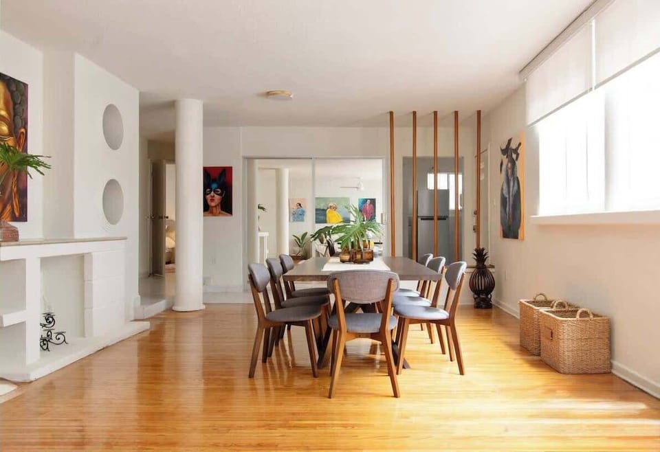Bright dining area with a wooden table, framed by large windows and polished wood floors. Artistic paintings and natural accents create a warm gallery-inspired setting for shared meals.