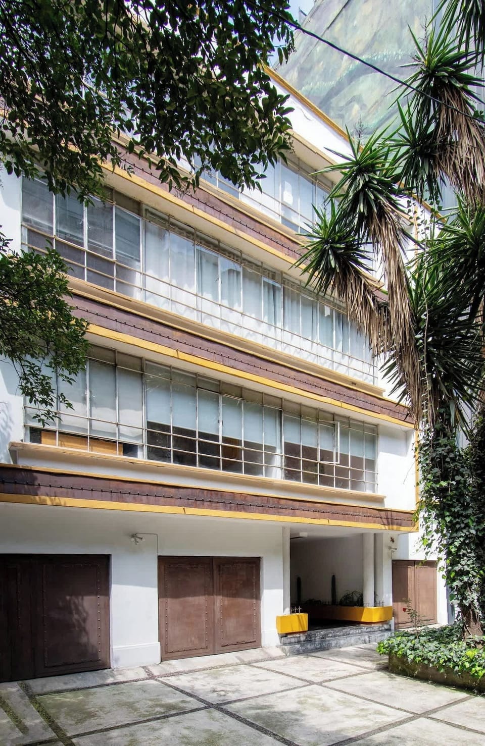 The building’s façade captures a true retro spirit, with wide glass windows, clean geometric lines, and mustard accents that recall mid-century design. Surrounded by tall palms and greenery, it blends urban character with timeless charm.