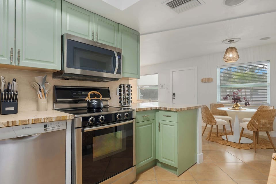 Bright, coastal kitchen with soft green cabinetry, stainless appliances, and spacious counters. Cook with ease, enjoy morning coffee, and gather nearby in the sunny dining nook—perfect for relaxed meals and beachy living.