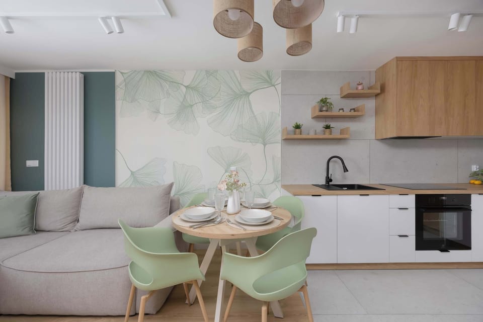 A dining area with a round table and pastel green chairs connected to the kitchen. The tropical wall design adds freshness to the space.