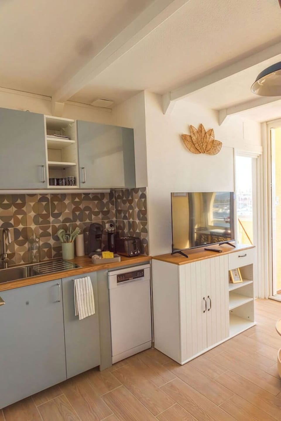 Cuisine équipée vue large – crédence carreaux rétro, plan de travail bois, TV murale et fenêtre lumineuse. Un espace ouvert et convivial pour préparer vos repas avec vue à Leucate.