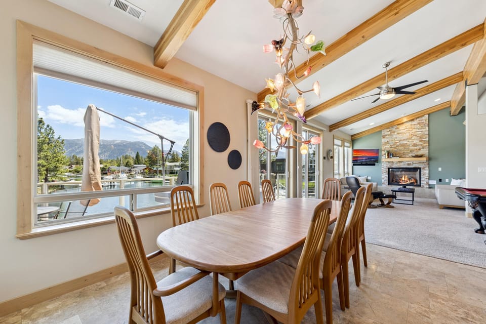 Light wood dining table seats eight beneath a glass floral chandelier, with large windows framing lake and mountain views.