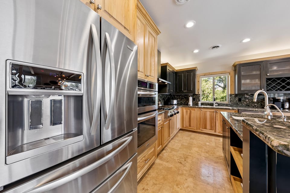 Stainless steel refrigerator and double oven stand beside granite counters and natural wood cabinets in the kitchen.