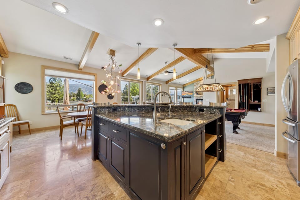 Black granite kitchen island with undermount sink stands on beige tile floors beneath wood ceiling beams and recessed lights.
