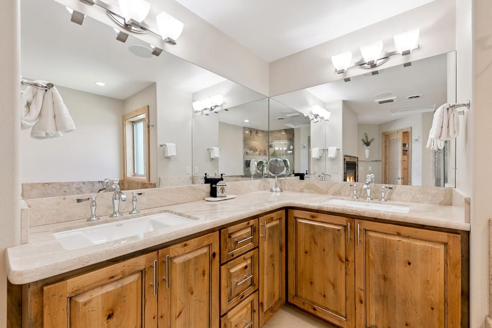 Bathroom vanity with dual sinks, polished stone countertops, natural wood cabinets, and large corner mirrors.