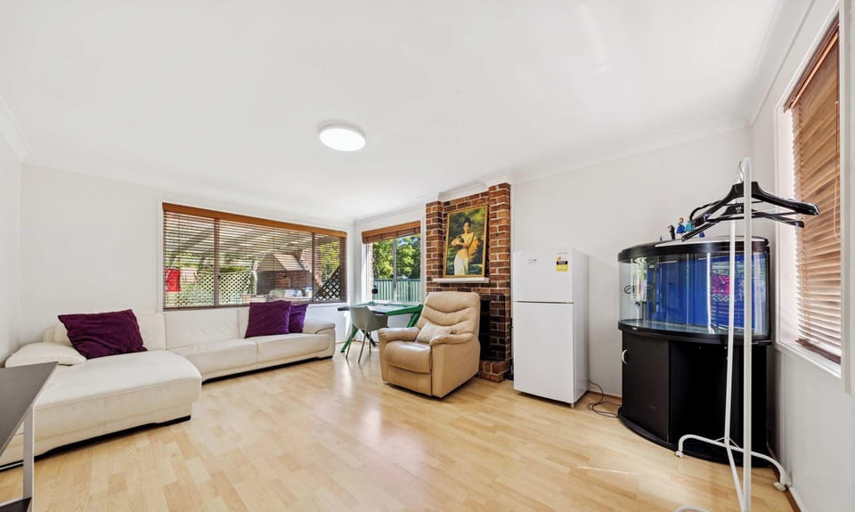 Spacious and light-filled living area featuring timber flooring, a white L-shaped sofa with accent cushions, and a beige recliner chair. The room includes a brick fireplace with artwork above, large windows with wooden blinds allowing natural light, 