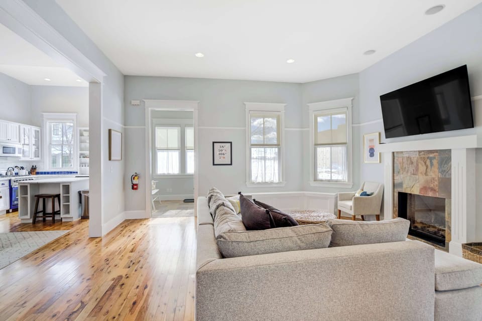 A bright living room with a stone fireplace, mounted Smart TV, and wraparound windows creates a relaxing space to gather after a day outdoors.