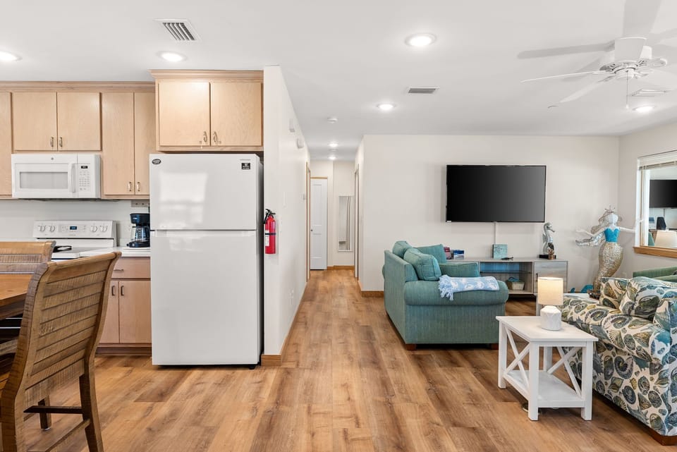 Your open-concept retreat flows seamlessly from kitchen to living area, with warm wood floors and coastal-inspired furnishings creating the perfect home base.