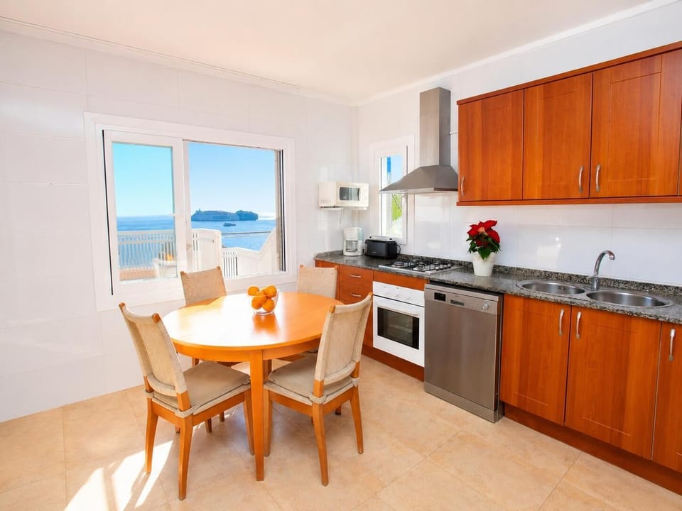 A fully equipped kitchen features warm wood cabinetry, modern appliances, and a circular dining table positioned next to a window with scenic sea views.