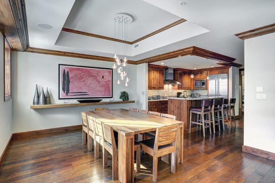 The spacious dining area features a long wooden table with stylish chairs, accented by modern pendant lights and a stunning pink artwork, all set against a backdrop of exquisite mountain views.