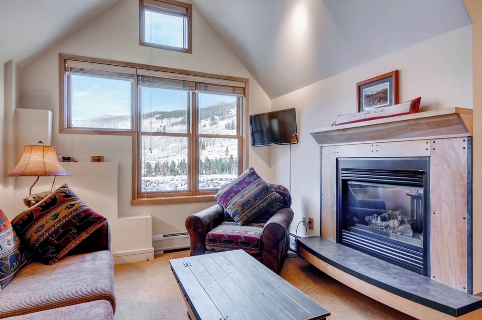 Warmth radiates from the elegant gas fireplace, perfectly complementing the cozy alpine atmosphere visible through large windows.