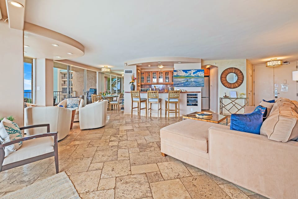 Spacious living room with cream furnishings and stone tile floors, sunlight spilling across glass doors while waves shimmer beyond.