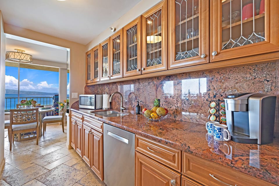 Wide kitchen view with granite counters and stainless appliances, sunlight streaming through windows as waves shimmer in the distance.