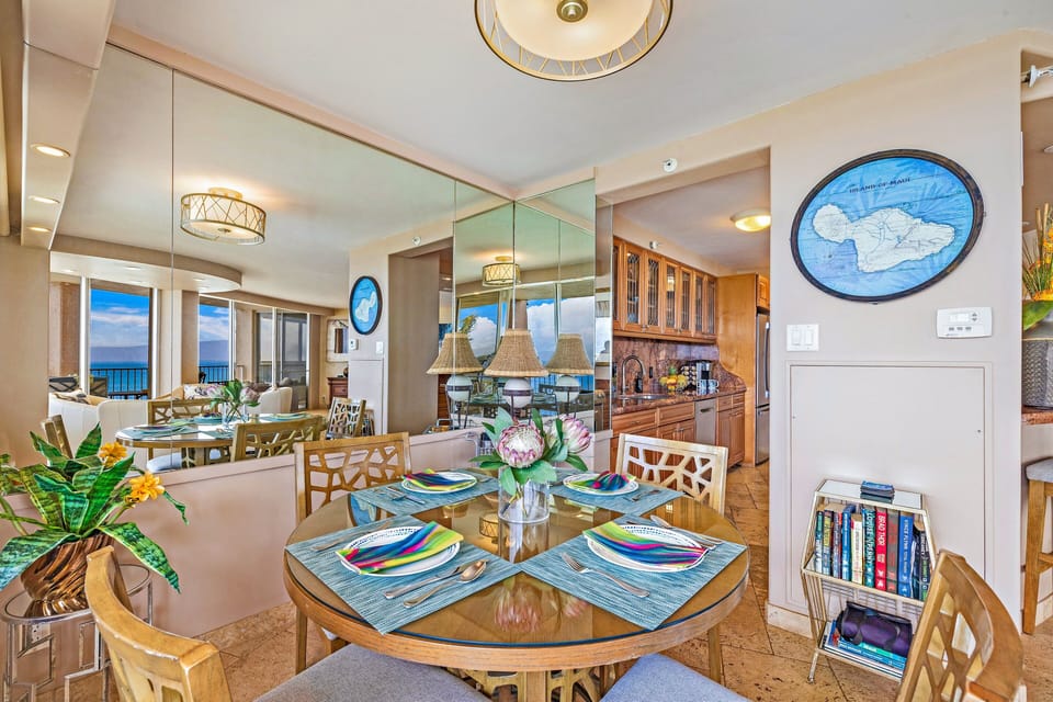 Round glass‑top table set with colorful placemats, mirrored walls reflecting ocean light while the scent of fresh flowers brightens the space.