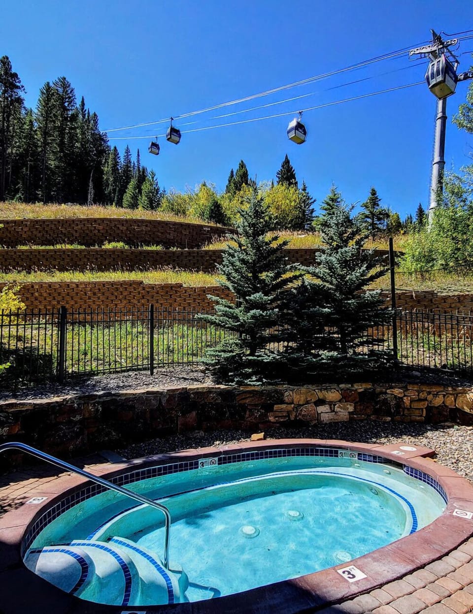 Hot tub surrounded by evergreen trees with mountain gondola overhead in a scenic alpine setting.