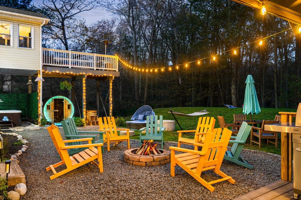 Outdoor space with Adirondack chairs, firepit, string lights, and woods.