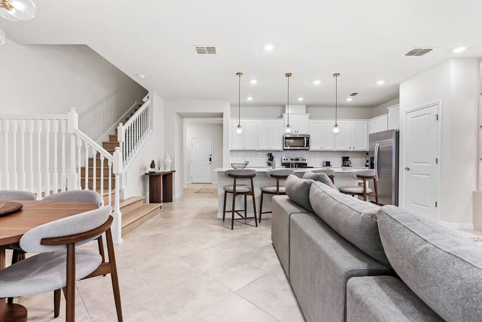 Experience a bright, airy living area with soft gray furnishings and warm wood accents, enhanced by elegant pendant lighting. The open layout seamlessly connects to a stylish kitchen, perfect for relaxing and entertaining.