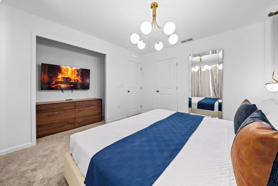 This cozy bedroom offers a plush queen bed with a rich blue blanket, soft neutral tones, and a stylish chandelier for a warm glow. Enjoy the modern convenience of a wall-mounted TV, perfect for a relaxing stay.