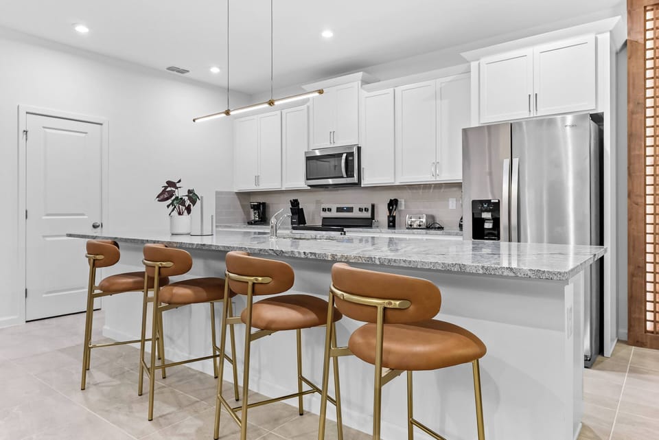 Welcome to a stylish kitchen with sleek white cabinetry and a spacious gray stone island. Comfortable leather bar stools and modern lighting create an inviting atmosphere, perfect for enjoying shared meals together.